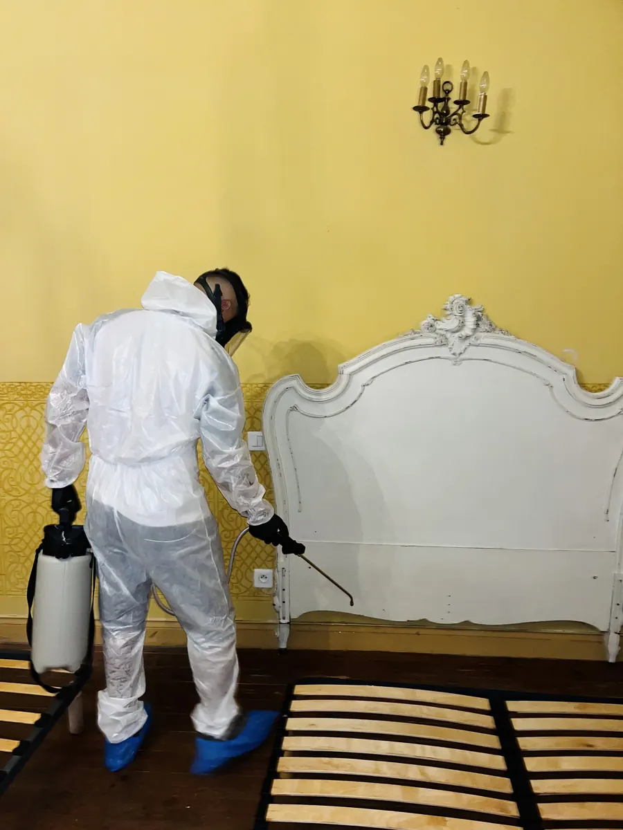 Technicien 3DTECH en combinaison, masque et surchaussures, pulvérisateur à la main, devant un lit démonté dans une chambre