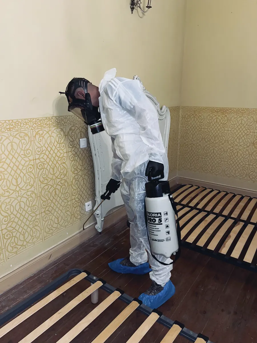 Technicien penché sur une tête de lit, pulvérisateur de cinq litres à la main, plinthes et lattes du sommier visibles